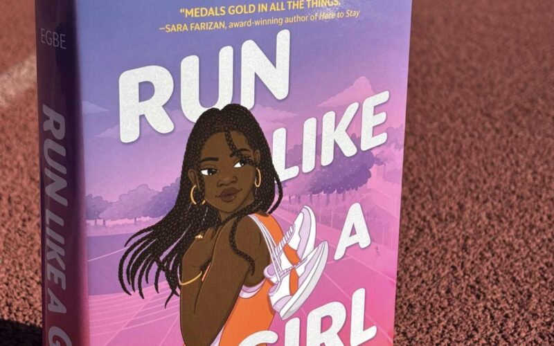 The book Run Like a Girl is sitting on a running track. A little bit of grass is in the far background. The cover of Run Like a Girl features a young Black woman with lots of braids that are a little past her shoulders. She is wearing gold hoops, bracelets and rings and is holding a pair white running shoes with an orange stripe over her shoulder. She's in an orange tank and orange shorts. She's looking over her shoulder with a slight smile. The background looks like a sunrise with yellow and orange at the bottom an pinks and purples up at the top.